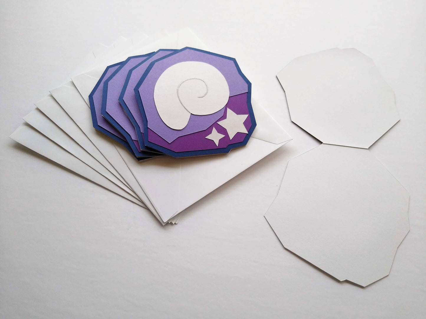 Four purple and white cards are stacked, splayed, on top of five white envelopes. The cards are designed to look like a shell and stars stuck inside a purple rock, like fossils. One card is opened up flat on the table beside them.