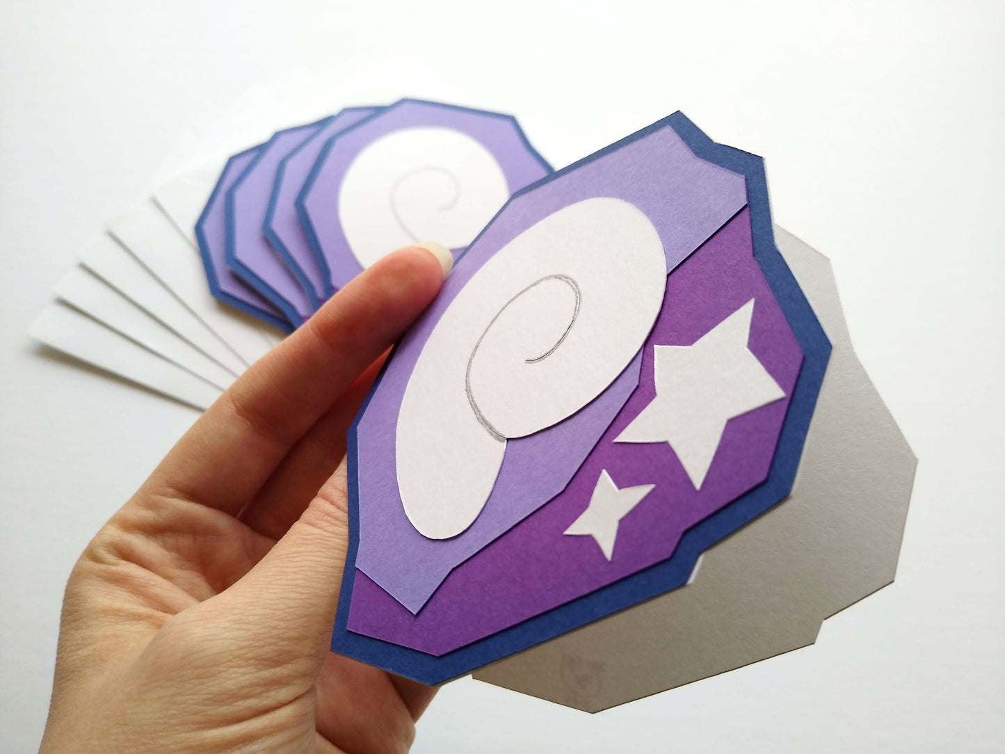 In the foreground is a hand holding a single card to the camera, opened slightly to show the white inside. The card is designed to look like a shell and two stars stuck inside a purple rock, like fossils. Four fossil cards are stacked on white envelopes in the background.