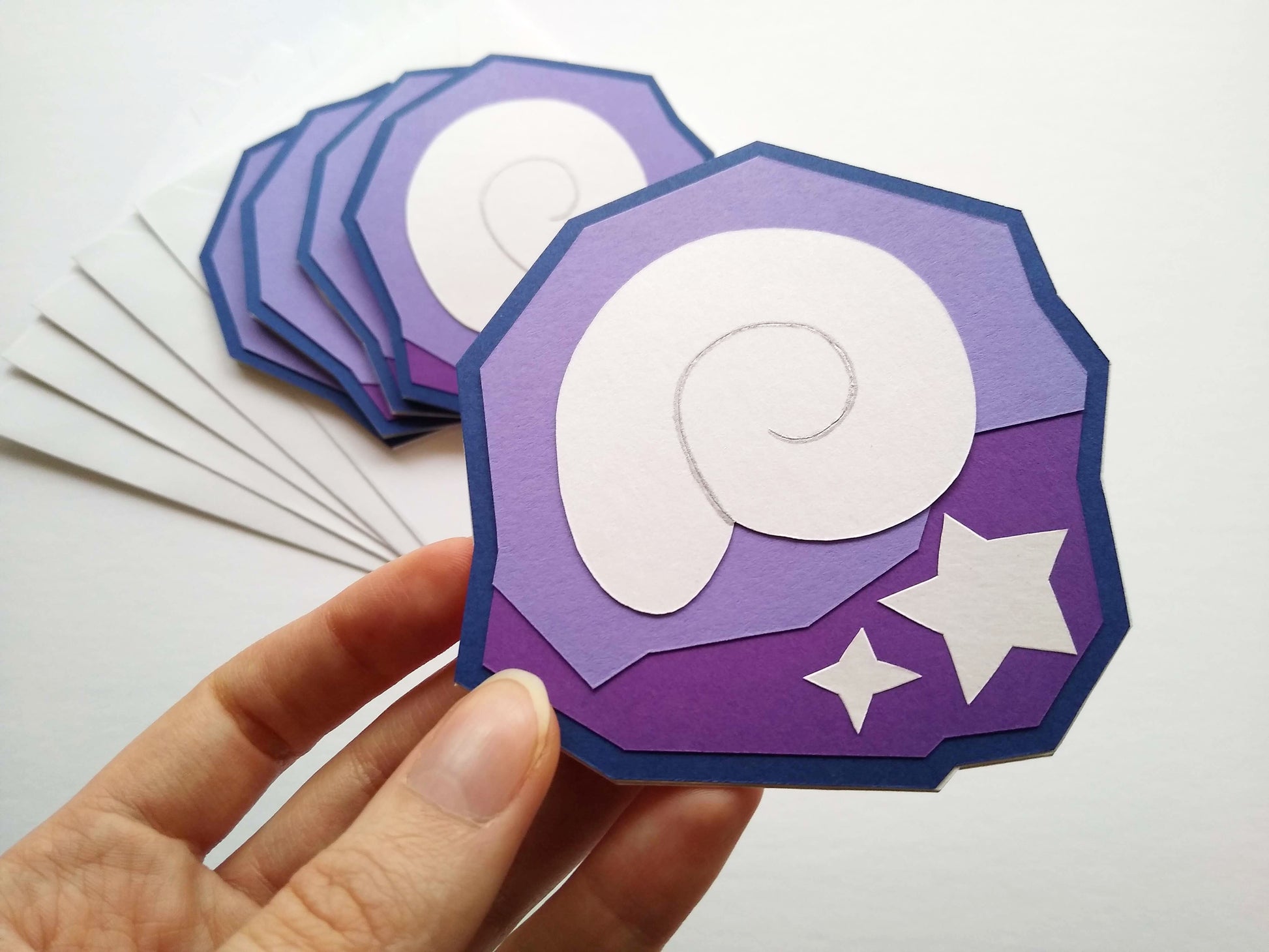 In the foreground is a hand holding a single card to the camera. The card is designed to look like a shell and two stars stuck inside a purple rock, like fossils. Four fossil cards are stacked on white envelopes in the background.