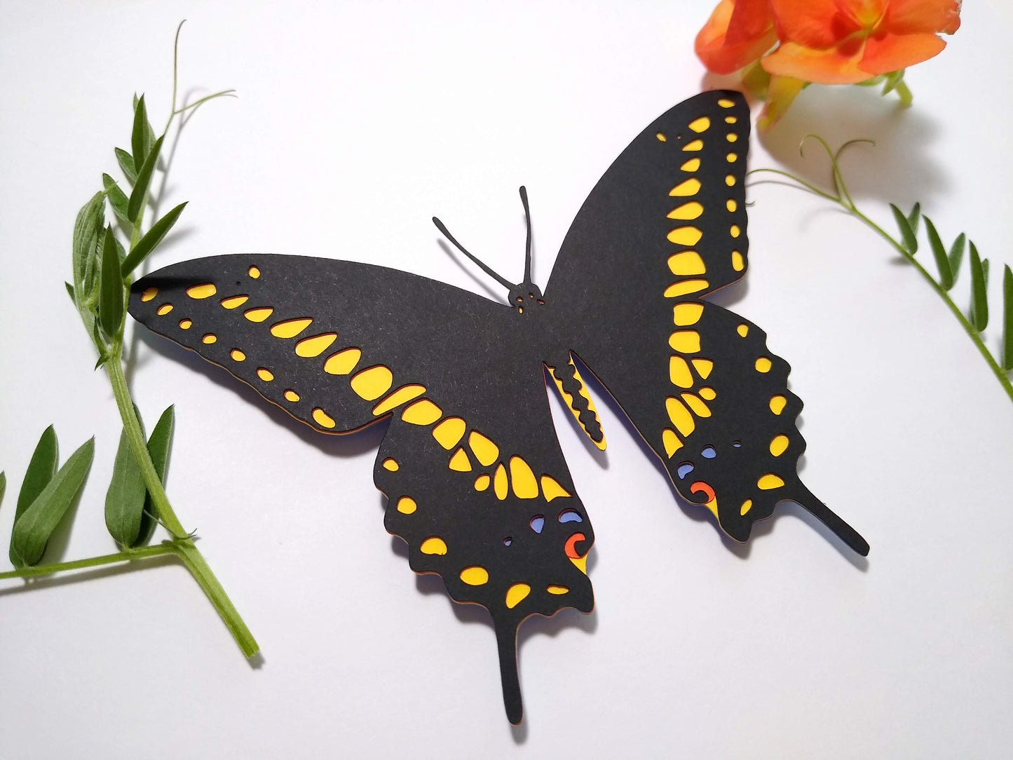Multi-layered paper butterfly, in the design of a Black Swallowtail butterfly. It rests next to several sprigs of leaves and a small orange flower.