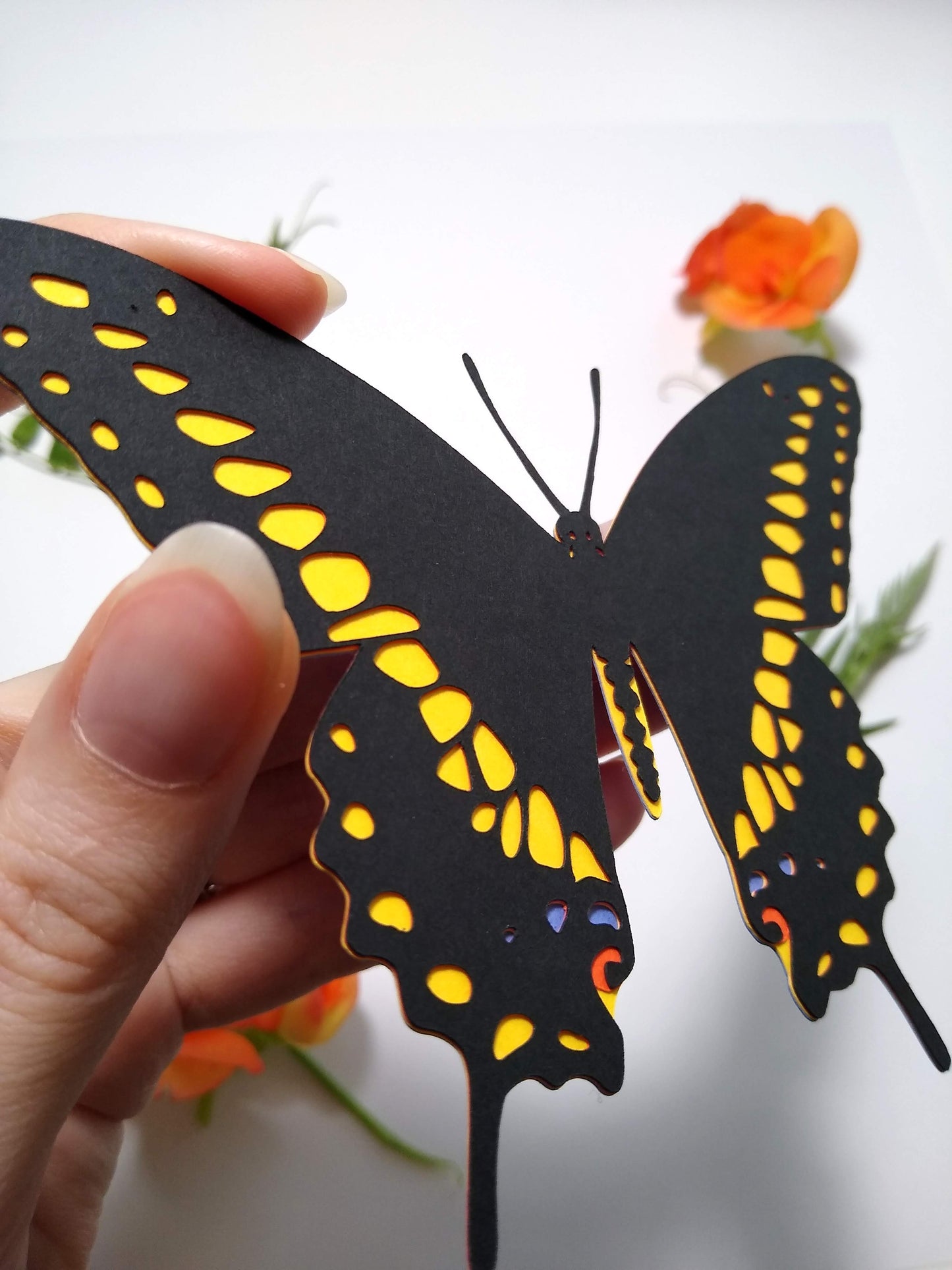 A hand holds a multi-layered paper butterfly to the camera, in the design of a Black Swallowtail butterfly. In the background are several sprigs of leaves and a small orange flower.