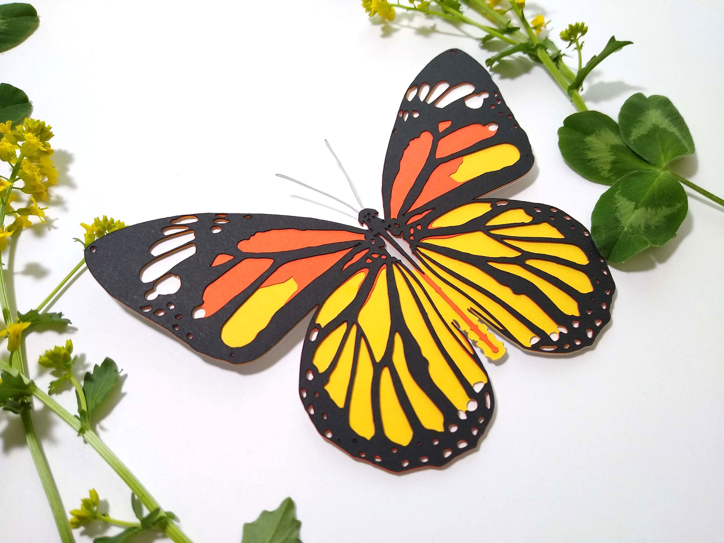 Multi-layered paper butterfly, in the design of a Common Tiger butterfly. It rests next to several sprigs of leaves and small yellow flowers.