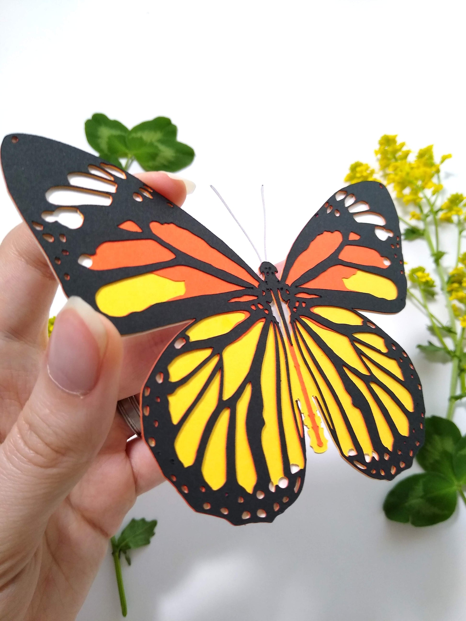 A hand holds a multi-layered paper butterfly to the camera, in the design of a Common Tiger butterfly. In the background are several sprigs of leaves and small yellow flowers.