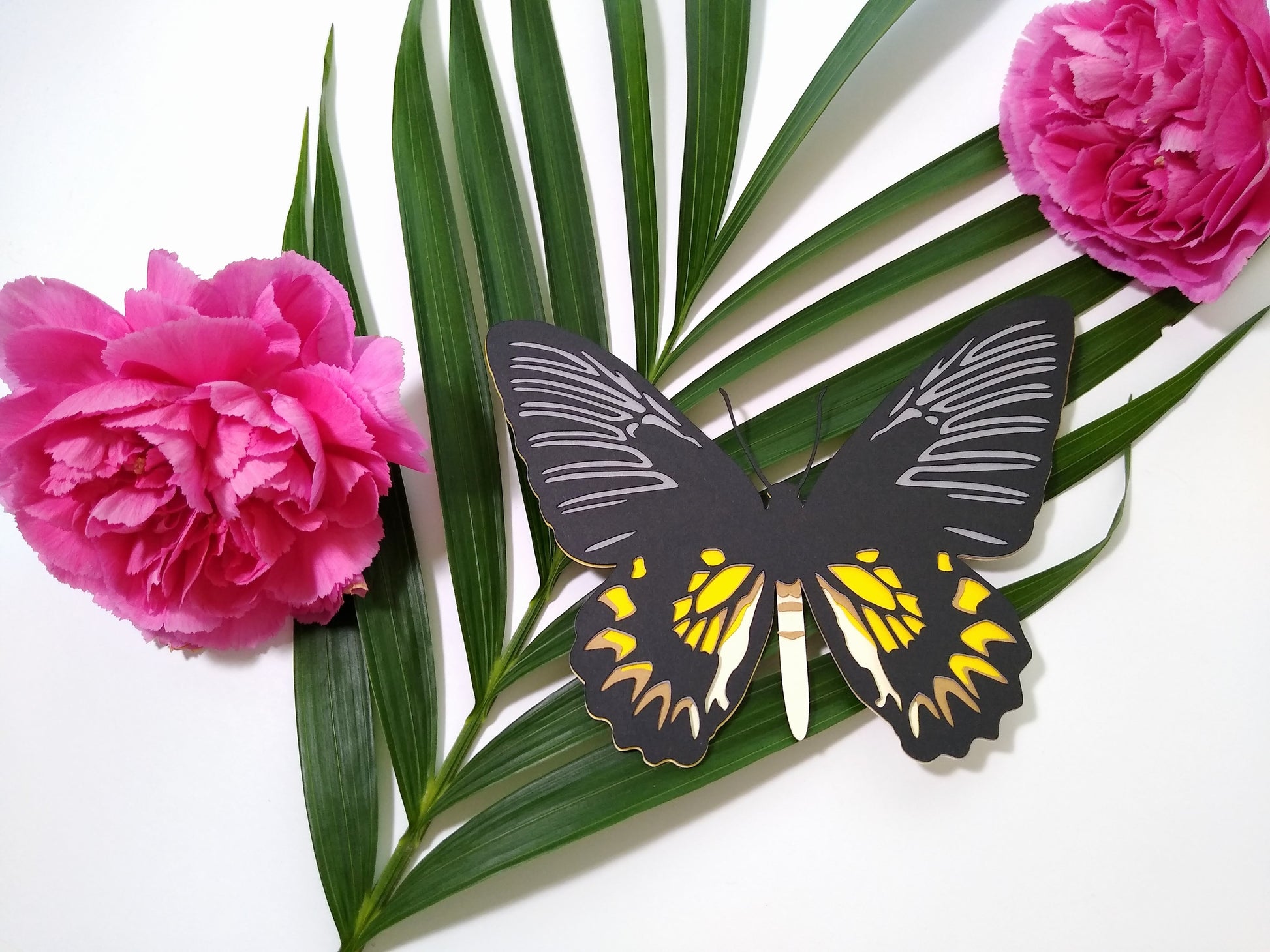 Multi-layered paper butterfly, in the design of a female Magellan Birdwing butterfly. It rests on a large sprig of leaves and beside two pink flowers.