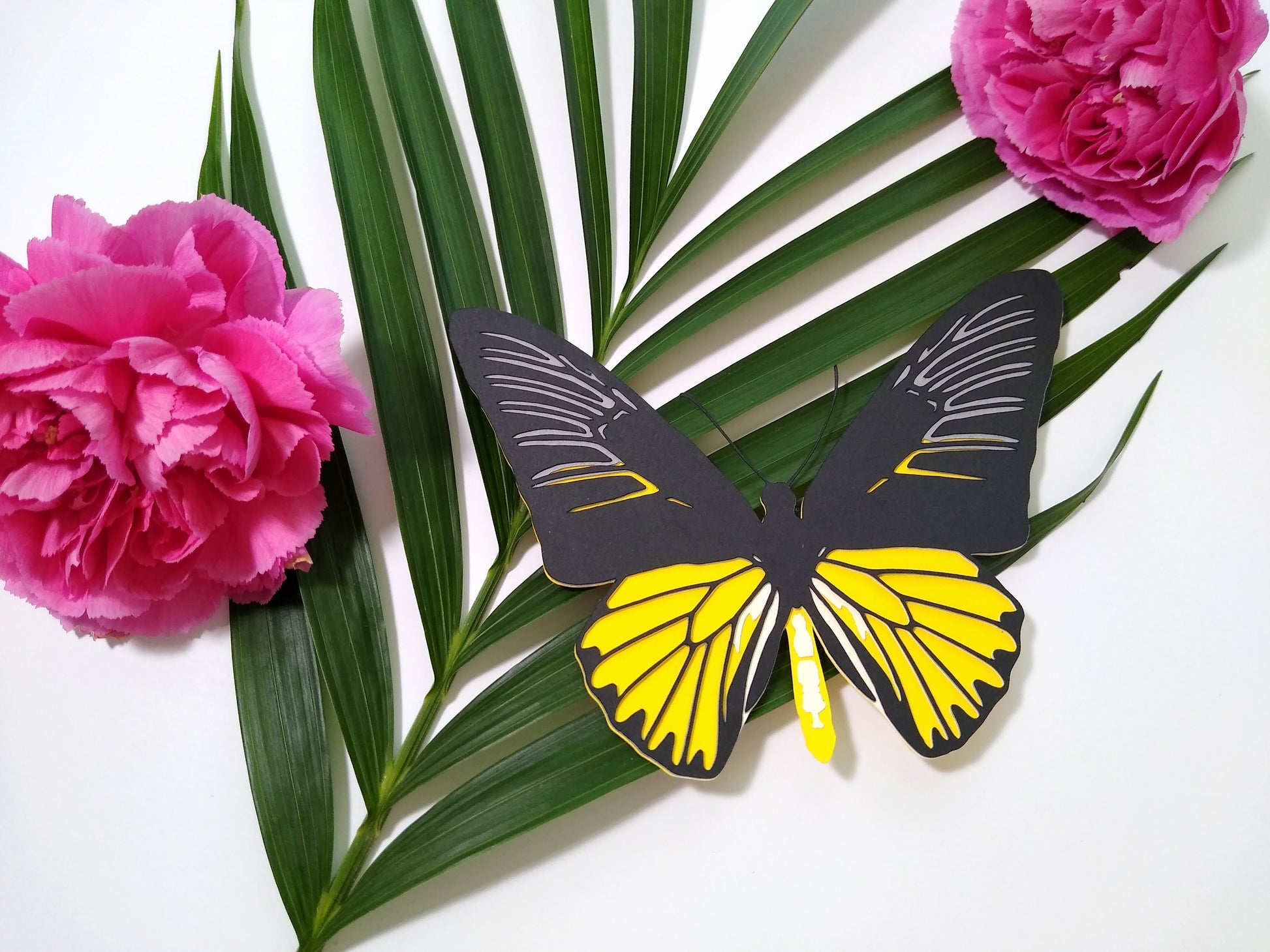 Multi-layered paper butterfly, in the design of a male Magellan Birdwing butterfly. It rests on a large sprig of leaves and beside two pink flowers.