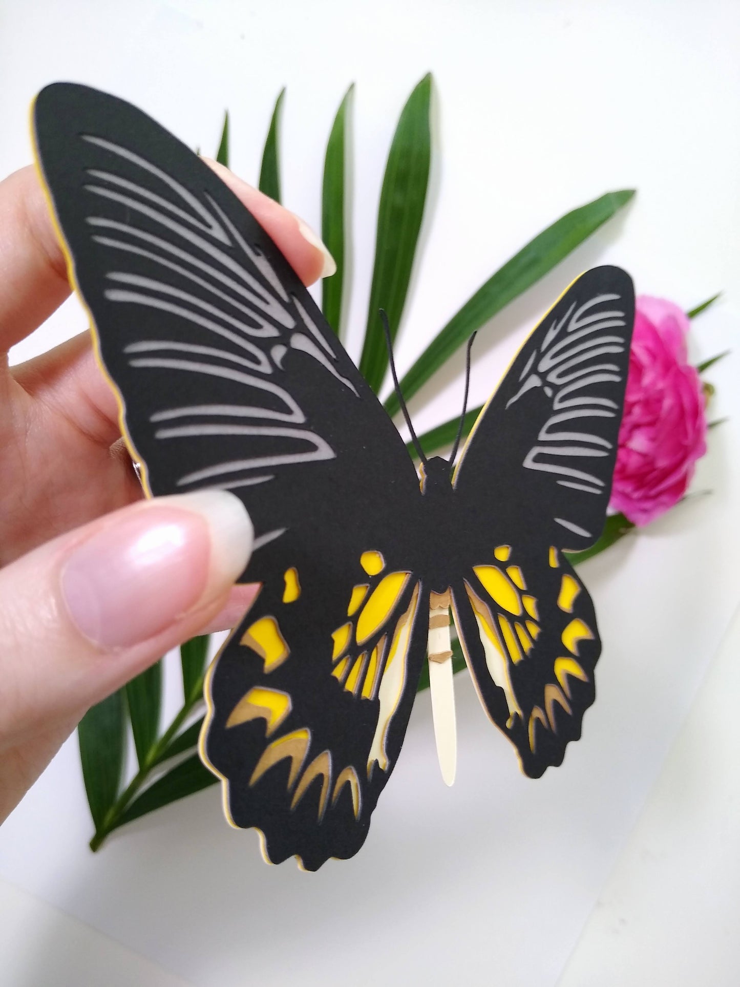 A hand holds a multi-layered paper butterfly to the camera, in the design of a female Magellan Birdwing butterfly. In the background are a large sprig of leaves and a pink flower.