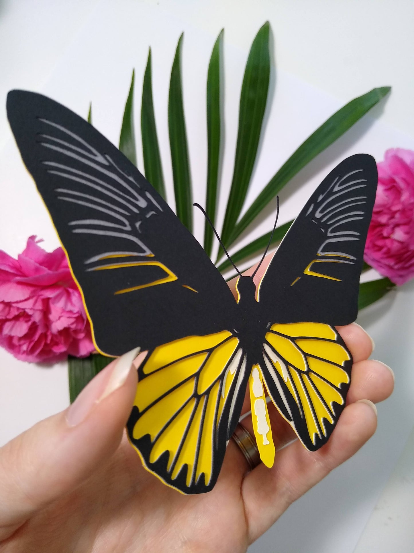 A hand holds a multi-layered paper butterfly to the camera, in the design of a male Magellan Birdwing butterfly. In the background are a large sprig of leaves and two pink flowers.