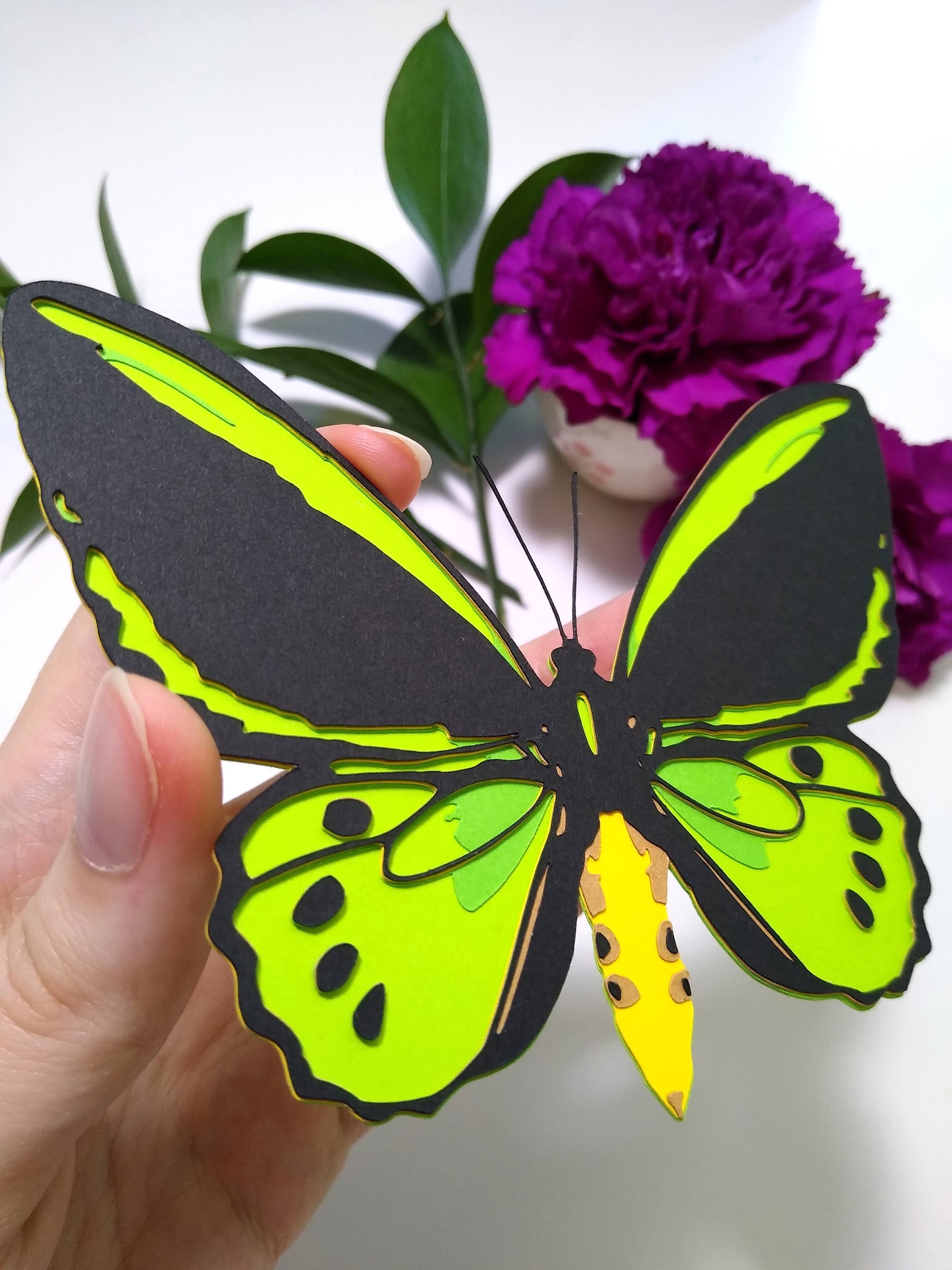 A hand holds a multi-layered paper butterfly to the camera, in the design of the front of a Cairns Birdwing butterfly. In the background are leaves and two purple flower.