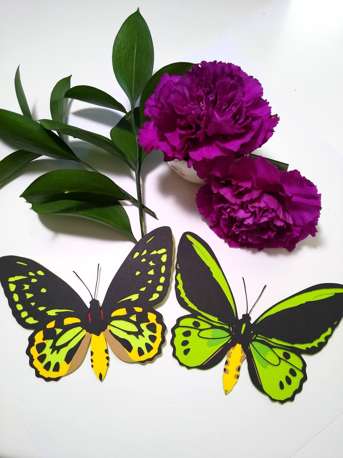 Two multi-layered paper butterflies rest on a table. They are in the design of the front and back of a Cairns Birdwing butterfly. They rest below sprigs of leaves and two purple flowers.