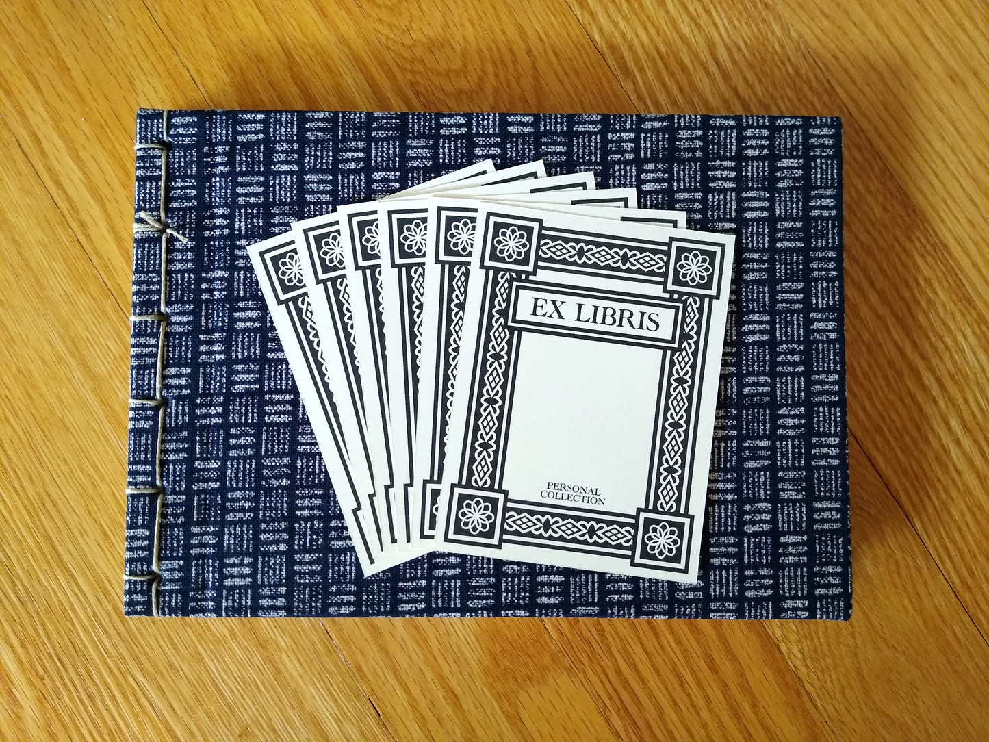 Six black and white bookplates rest on top of a handbound journal with cream and blue hash marks. The bookplates say Ex Libris and Personal Collection, and have a thick art deco border around the edges.