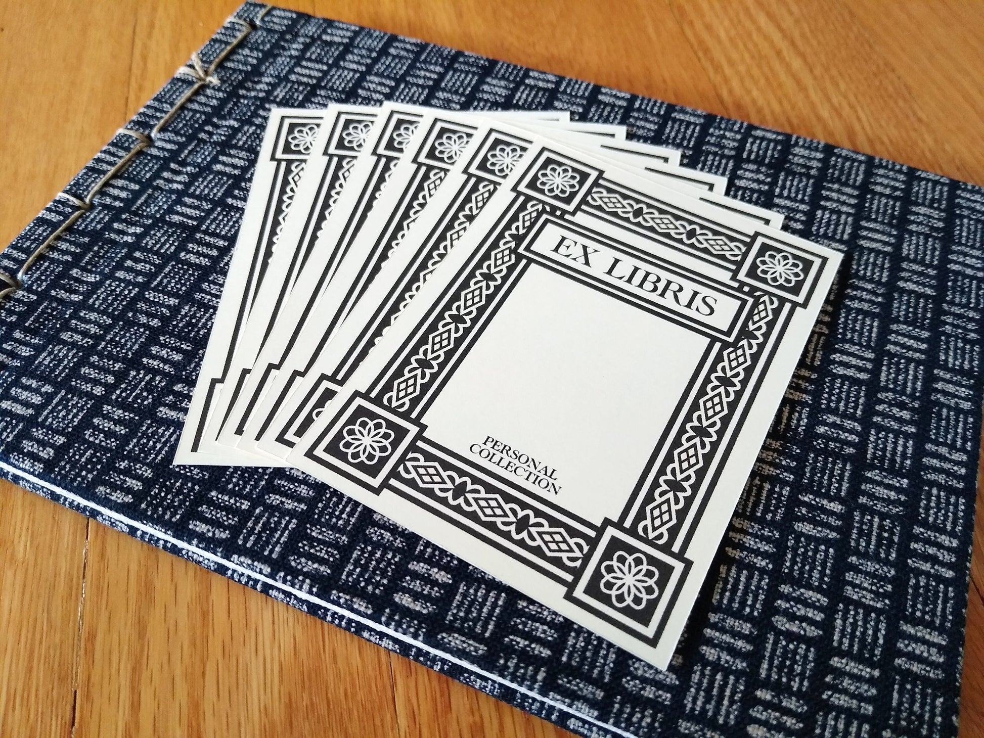 Six black and white bookplates rest on top of a handbound journal with cream and blue hash marks. The bookplates say Ex Libris and Personal Collection, and have a thick art deco border around the edges.