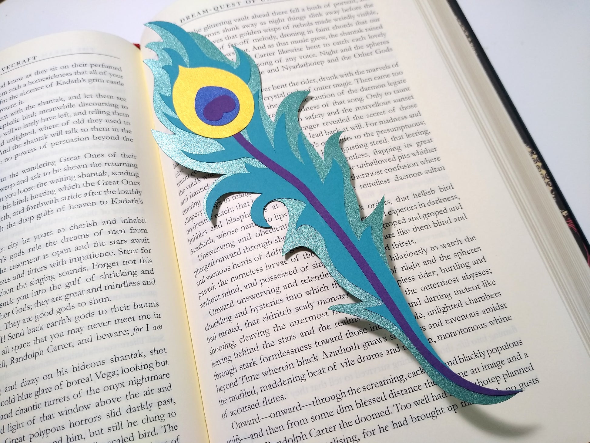 A multi-layer paper bookmark lays on an open book. Designed to look like a long peacock feather, the layers include a shiny teal, gold, purple, metallic blue, and a flat teal.