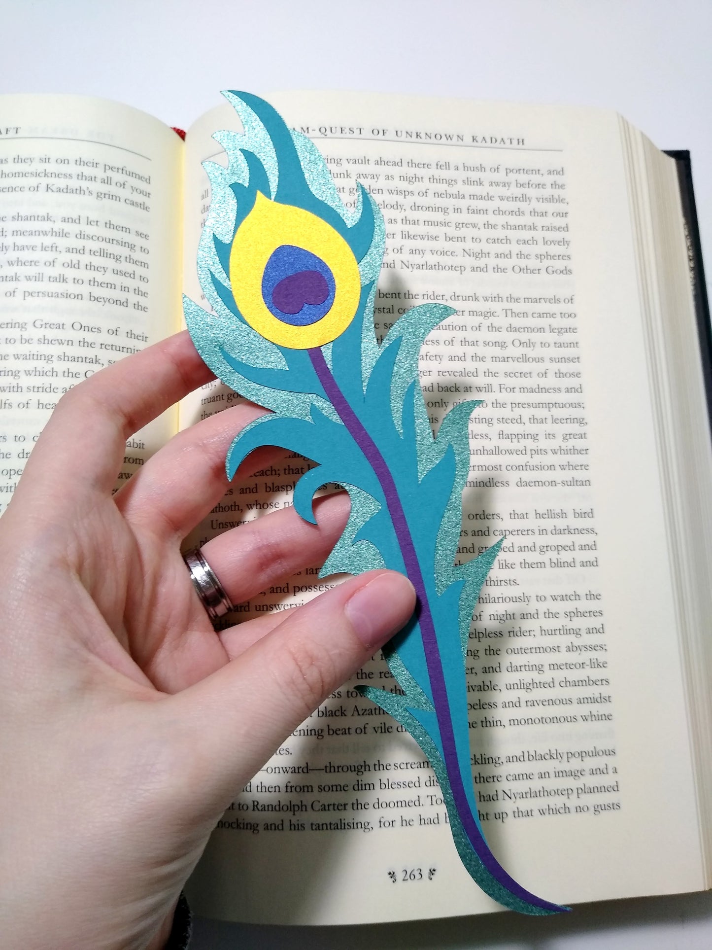A hand holds a multi-layer paper bookmark over top of an open book. Designed to look like a long peacock feather, the layers include a shiny teal, gold, purple, metallic blue, and a flat teal.