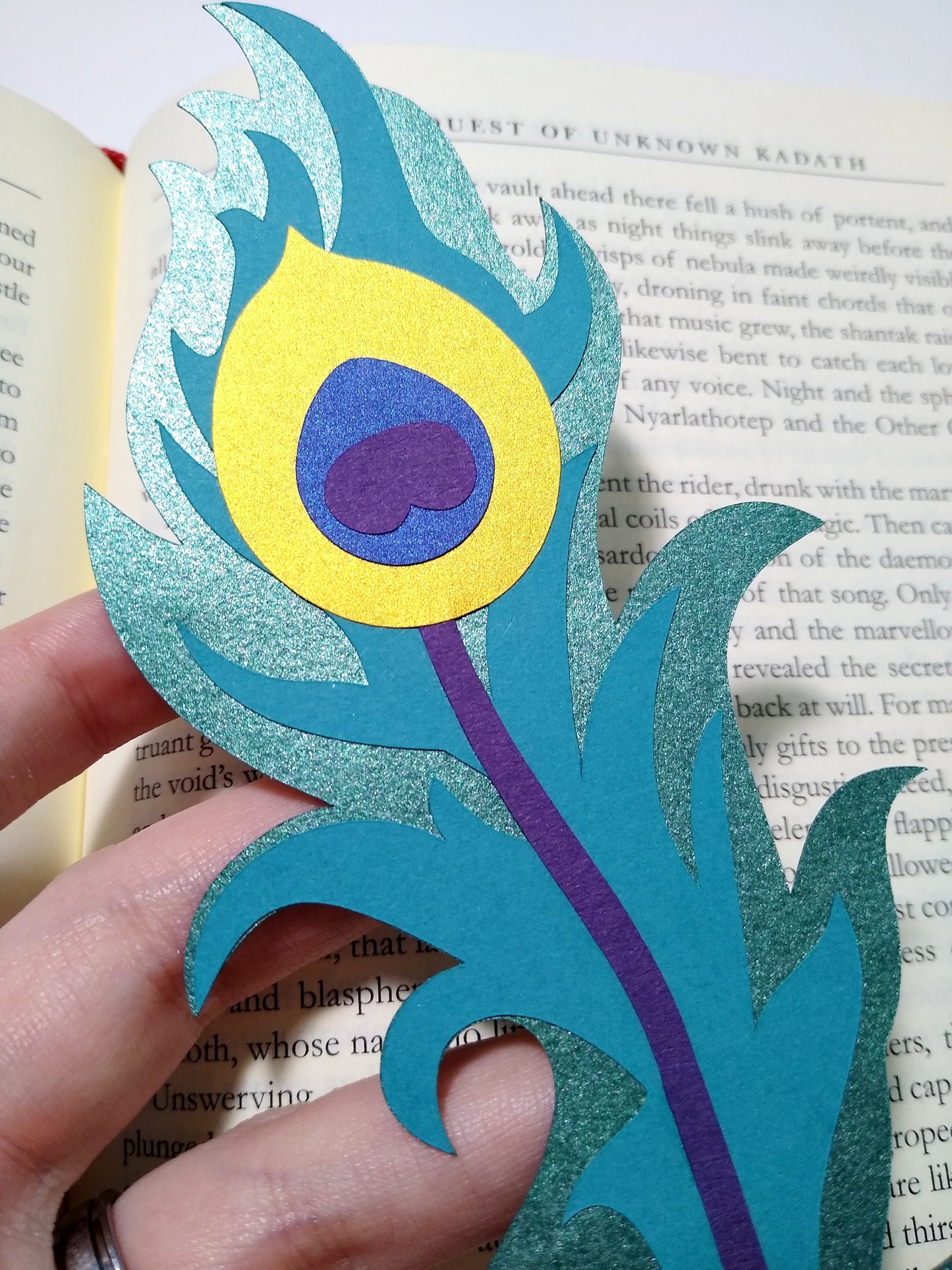 A close up of a multi-layer paper bookmark held over top of an open book. Designed to look like a long peacock feather, the layers include a shiny teal, gold, purple, metallic blue, and a flat teal.