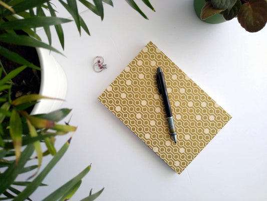 A handmade journal laying on a white desk beside two potted plants and a set of rings. A black mechanical pencil rests on top. The cover of the journal is cream with gold hexagons.