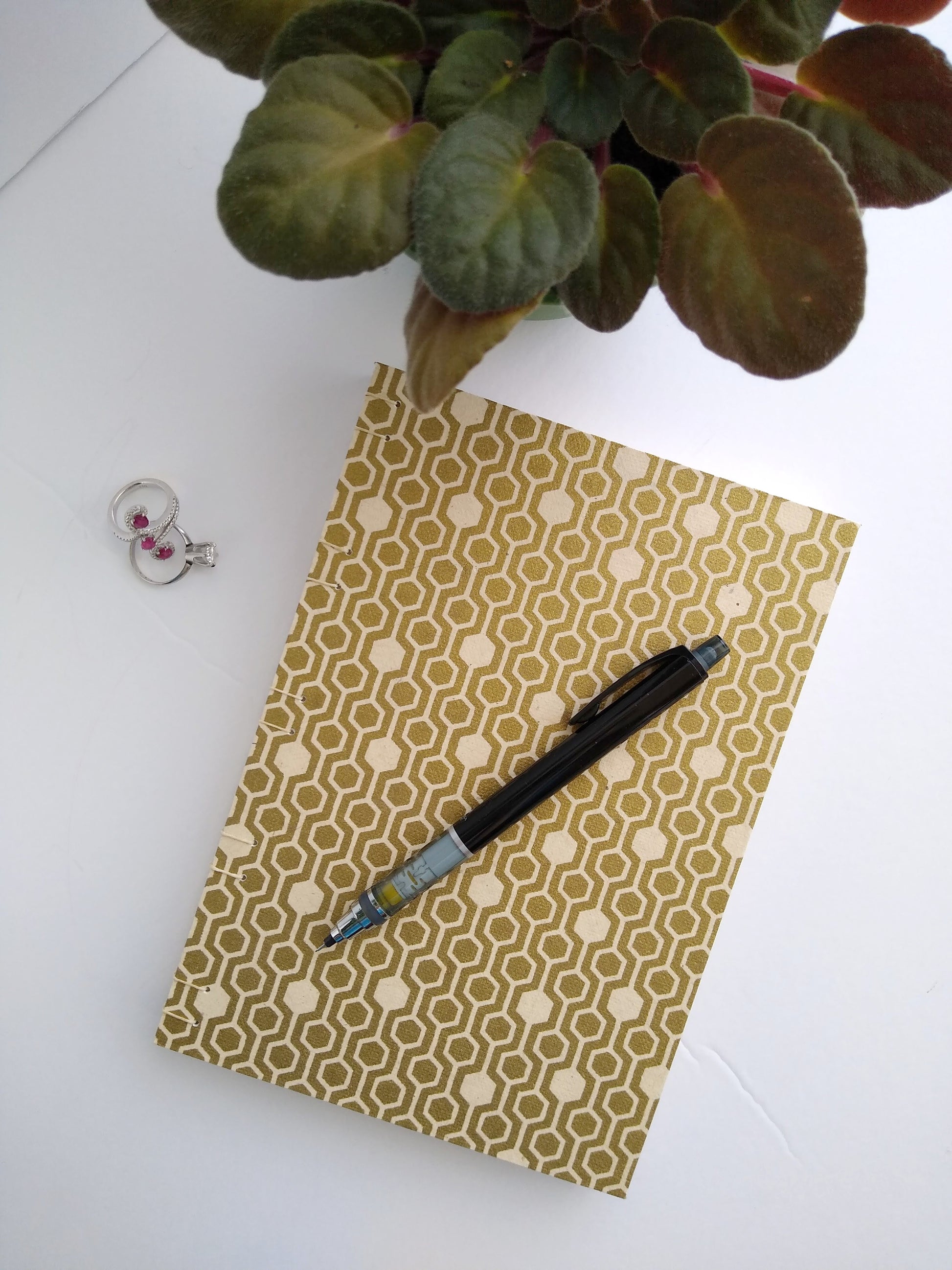 A handmade journal laying on a white desk beside a potted plant and a set of rings. A black mechanical pencil rests on top. The cover of the journal is cream with gold hexagons.