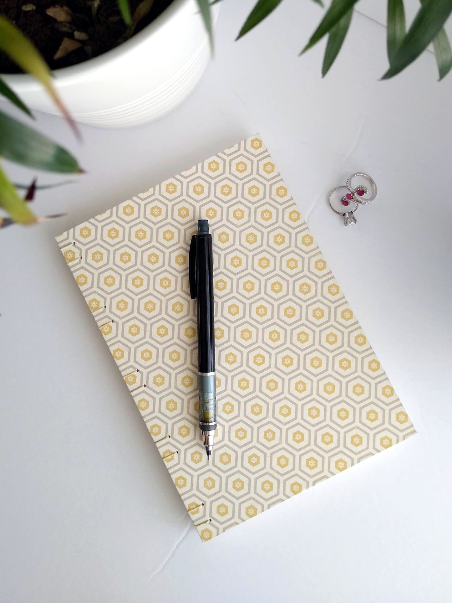 A handmade journal laying on a white background beside a potted plant and a set of rings with a black mechanical pencil resting on top. The cover of the journal is cream with concentric hexagons all over it in grey and yellow. 