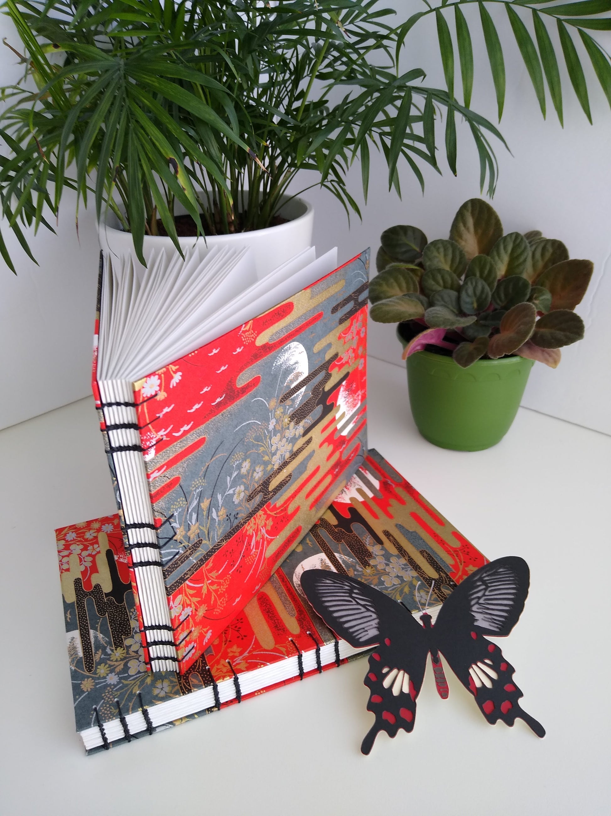 Two handmade journals are positioned beside two potted plants and a paper butterfly. One is laying flat and the other is standing upright on top of it. The cover of the journals are complete scenes with red and grey skies with partial moon outlines, flowers, birds, and gold and black stylized clouds. The journals are angled to show their open spines and black thread stitching them together.