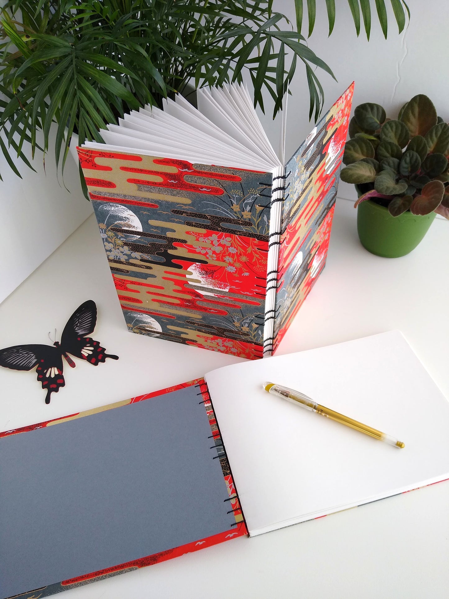 Two handmade journals are positioned beside two potted plants and a paper butterfly. One is laying flat and the other is standing upright. The one laying flat is opened, showing the grey endpaper and has a gold pen laying on the blank white pages. The cover of the journals are complete scenes with red and grey skies with partial moon outlines, flowers, birds, and gold and black stylized clouds. The upright journal is angled to show its open spine and black thread stitching it together.