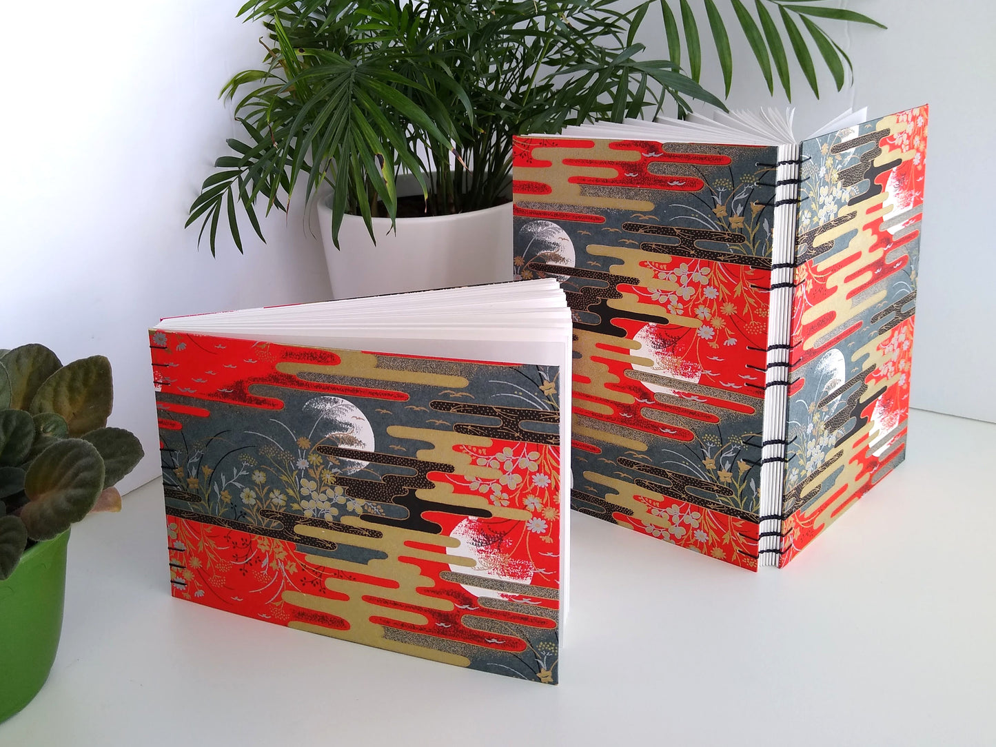 Two handmade journals are standing upright beside two potted plants. The covers of the journals are complete scenes with red and grey skies with partial moon outlines, flowers, birds, and gold and black stylized clouds. One journal is angled to show its open spine and black thread stitching it together.