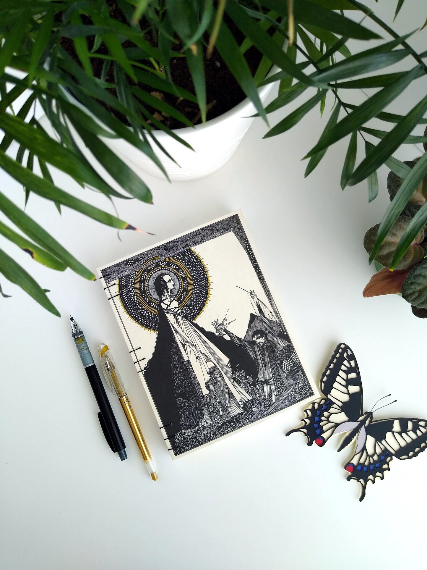 A handmade journal laying on a white desk beside two potted plants, a paper butterfly, a gold pen, and a black mechanical pencil. The cover of the journal is cream with printed with an illustration by Harry Clarke. It has a woman standing with a stylized halo/starburst behind her head with a man in supplication at her feet.