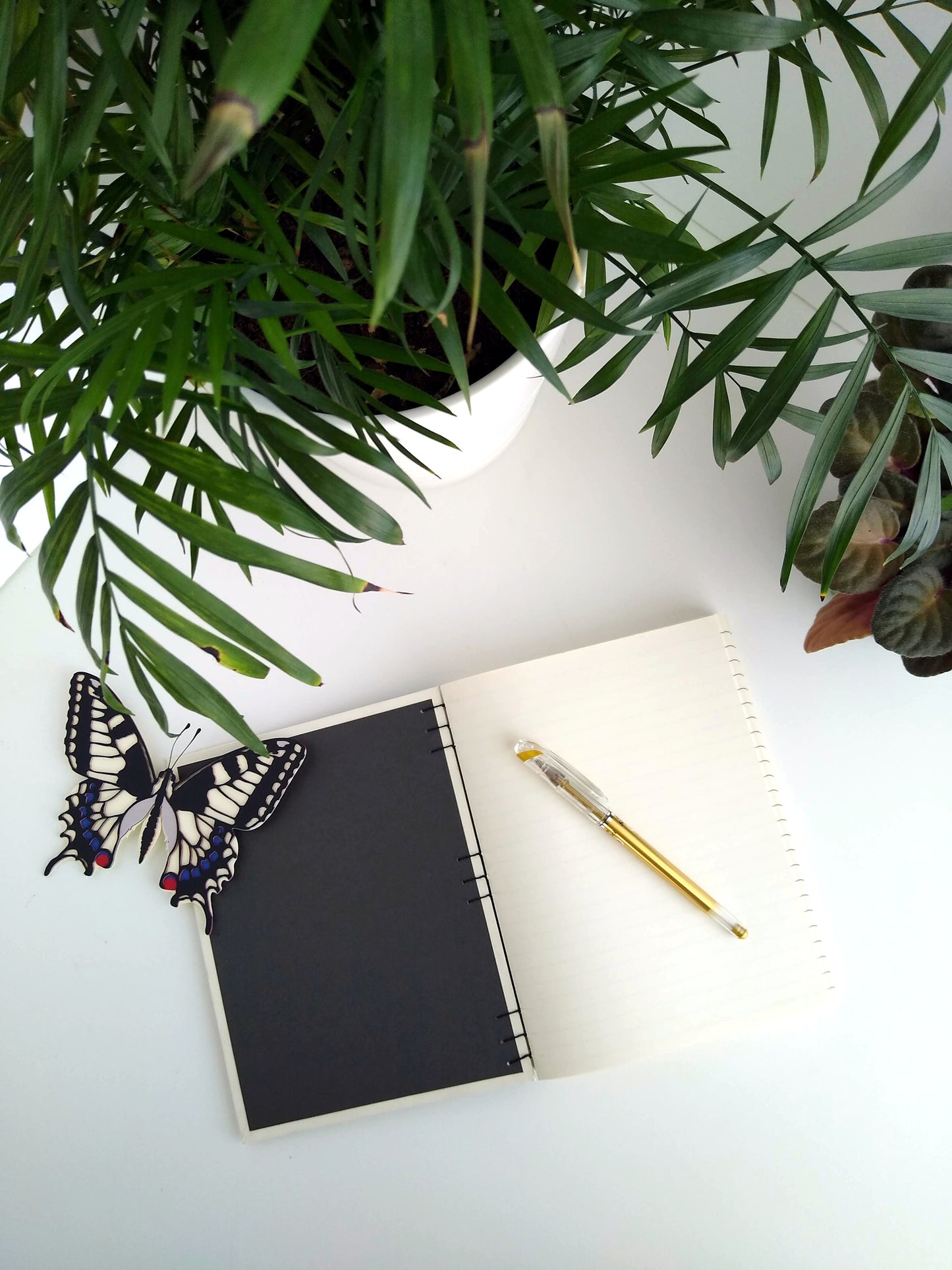 An open journal lays on a white desk next to two potted plants and a paper butterfly. The journal has a grey thread connecting the pages at the spine, dark grey endpapers and a blank, cream first sheet. A gold pen rests on top of the pages.