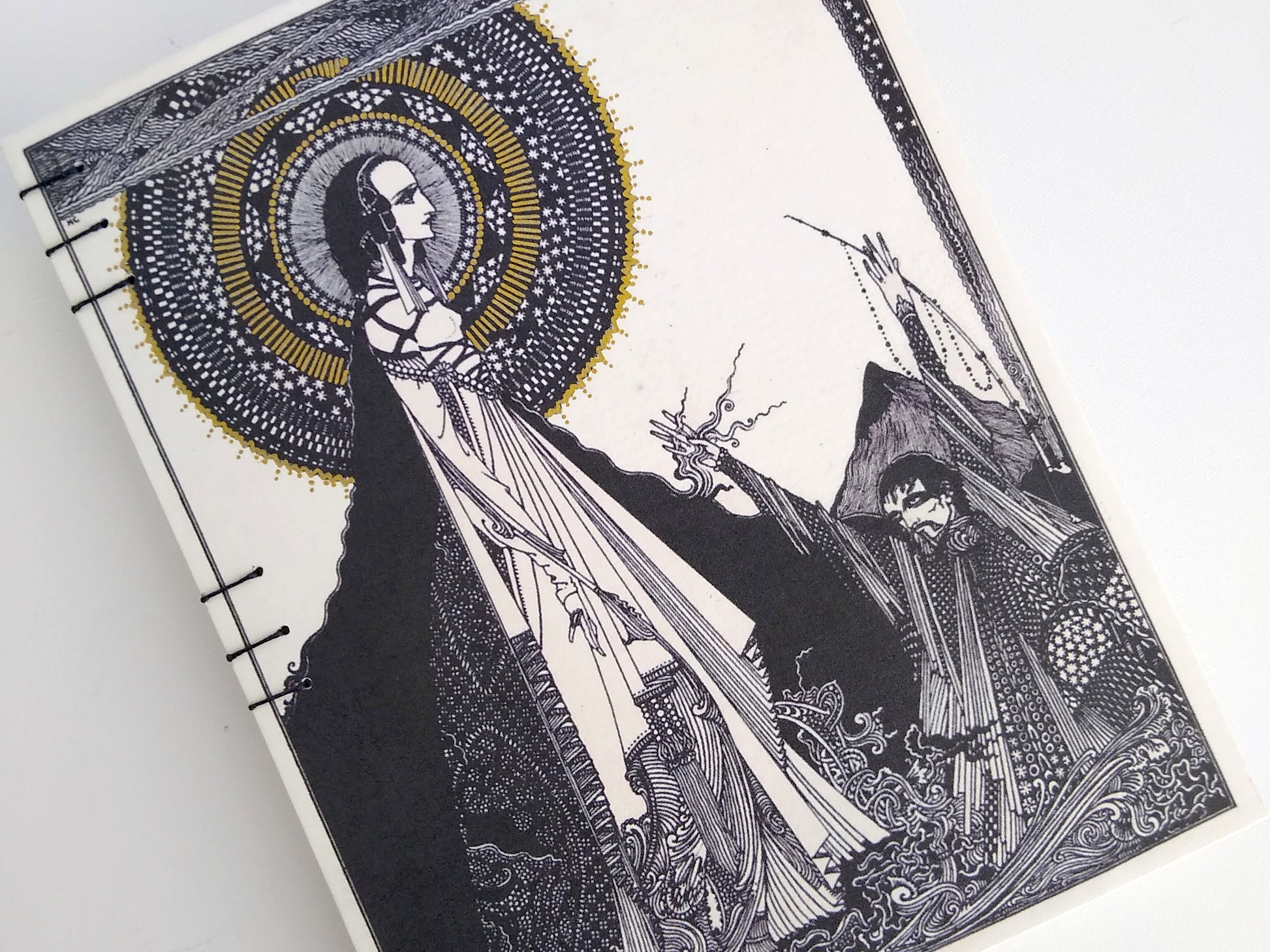 A closeup of a handmade journal on a white background. The cover of the journal is cream with printed with an illustration by Harry Clarke. It has a woman standing with a stylized halo/starburst behind her head with a man in supplication at her feet.