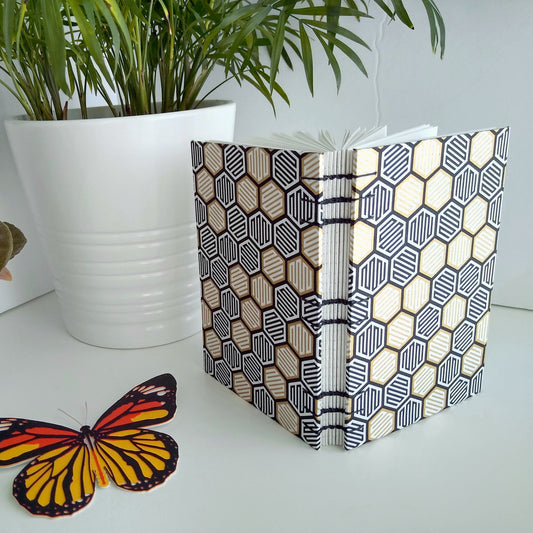 Handmade, open spine journal with abstract gold, white, and black hexagons over the covers. It's opened and standing upright next to a potted plant and paper butterfly.
