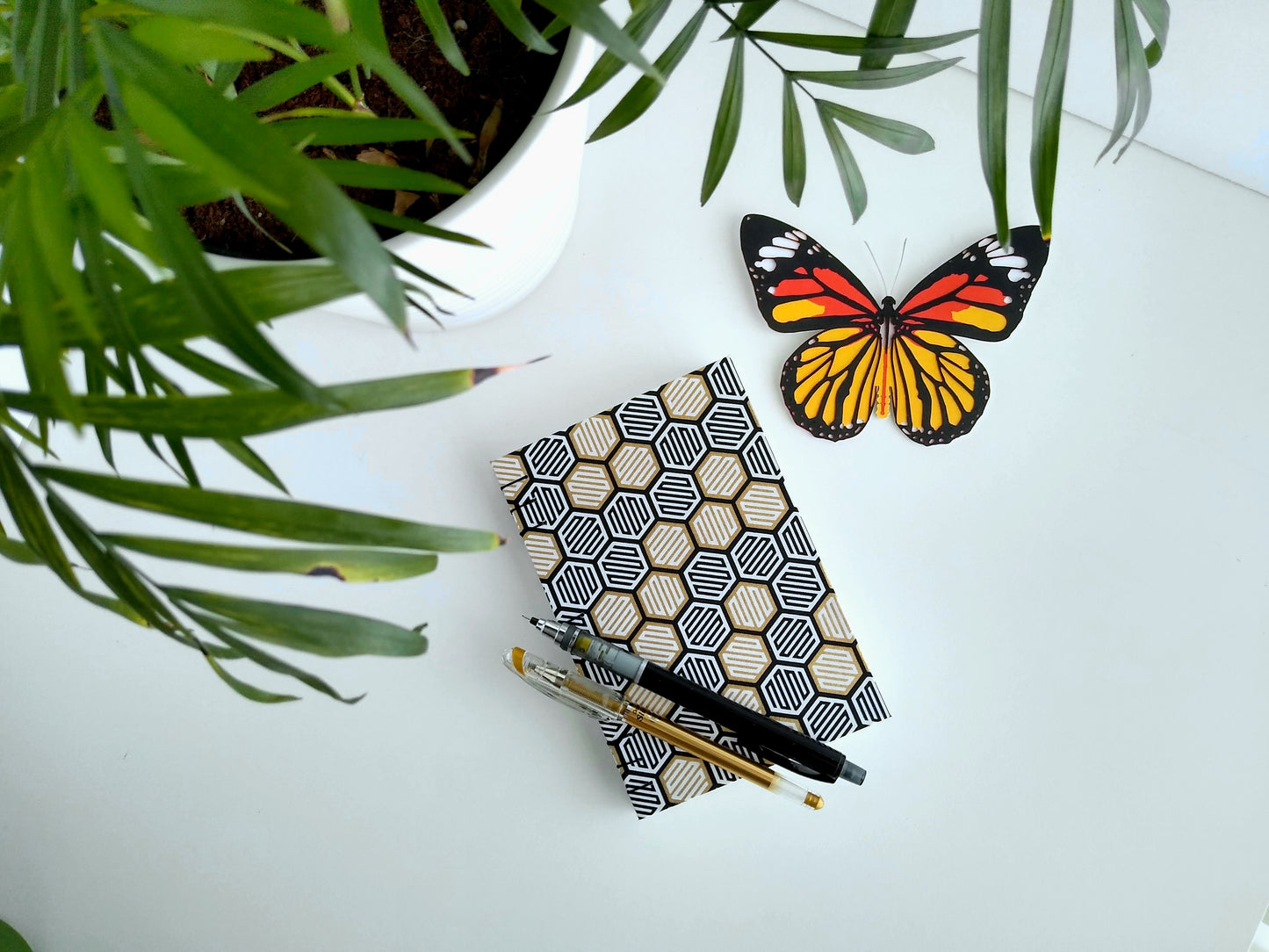 Handmade journal with abstract gold, white, and black hexagons over the cover. It's laying on a desk next to a potted plant and paper butterfly, with a gold pen and black mechanical pencil resting on top of it.