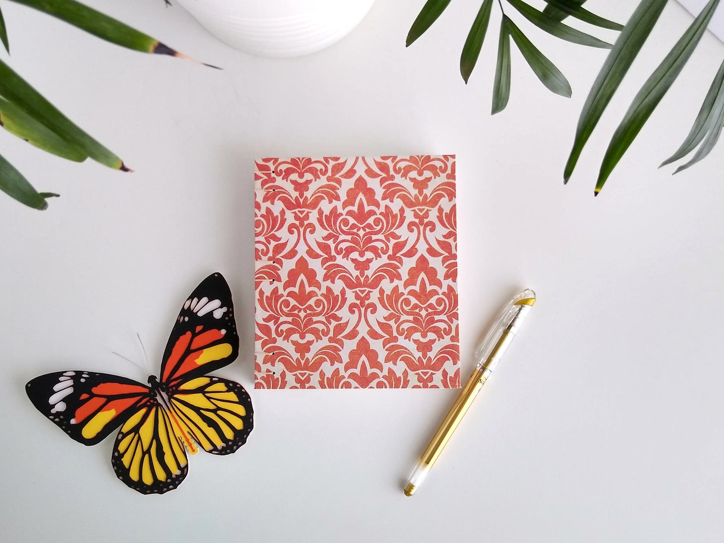 A handmade journal lays on a white desk beside a potted plant, a gold pen, and a paper butterfly. The cover of the journal is cream with an orange damask pattern printed across it.