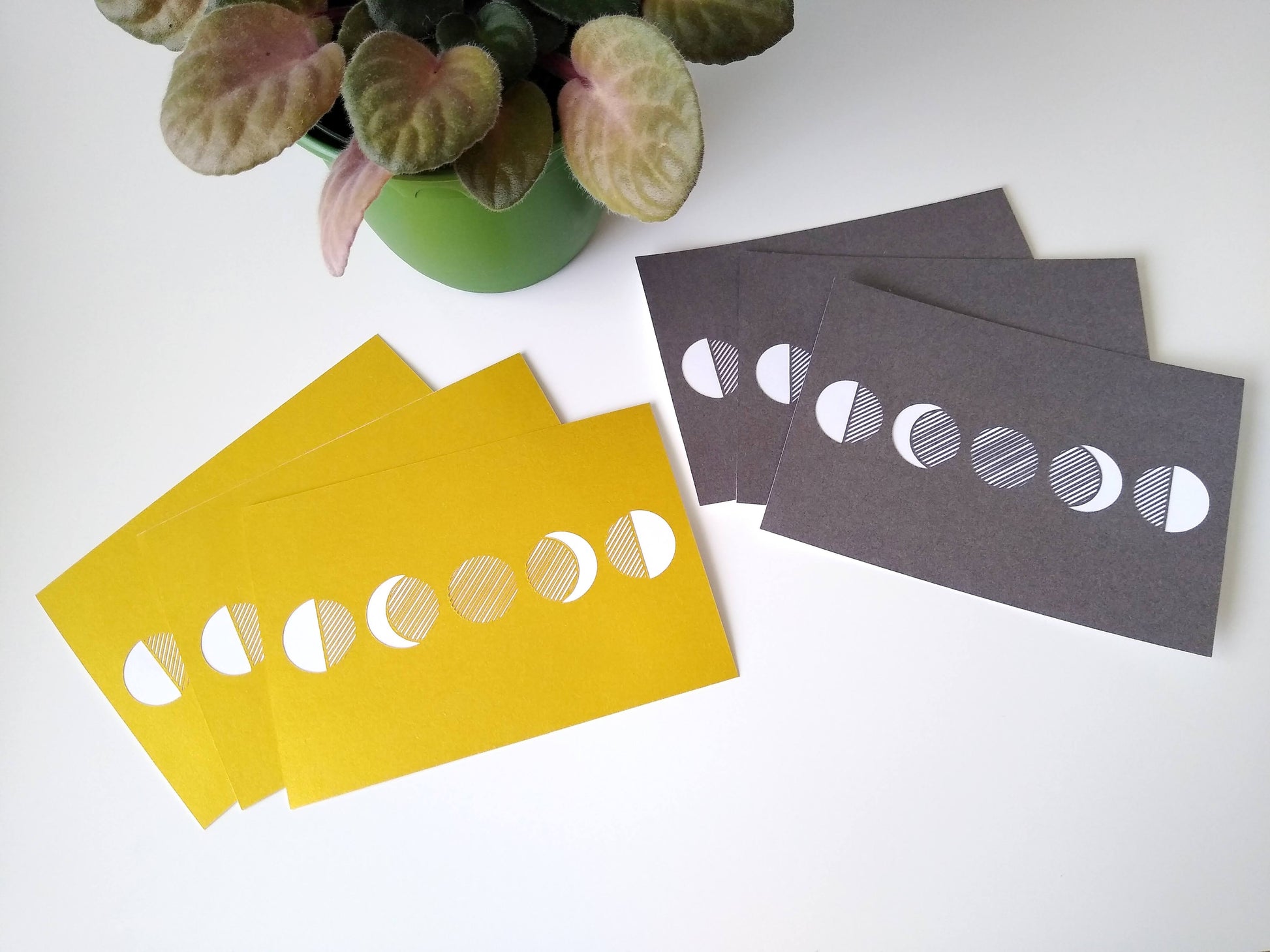Two groups of three postcards rest on a table beside a potted plant. One is metallic grey and the other is gold, both with five moon phases on them.