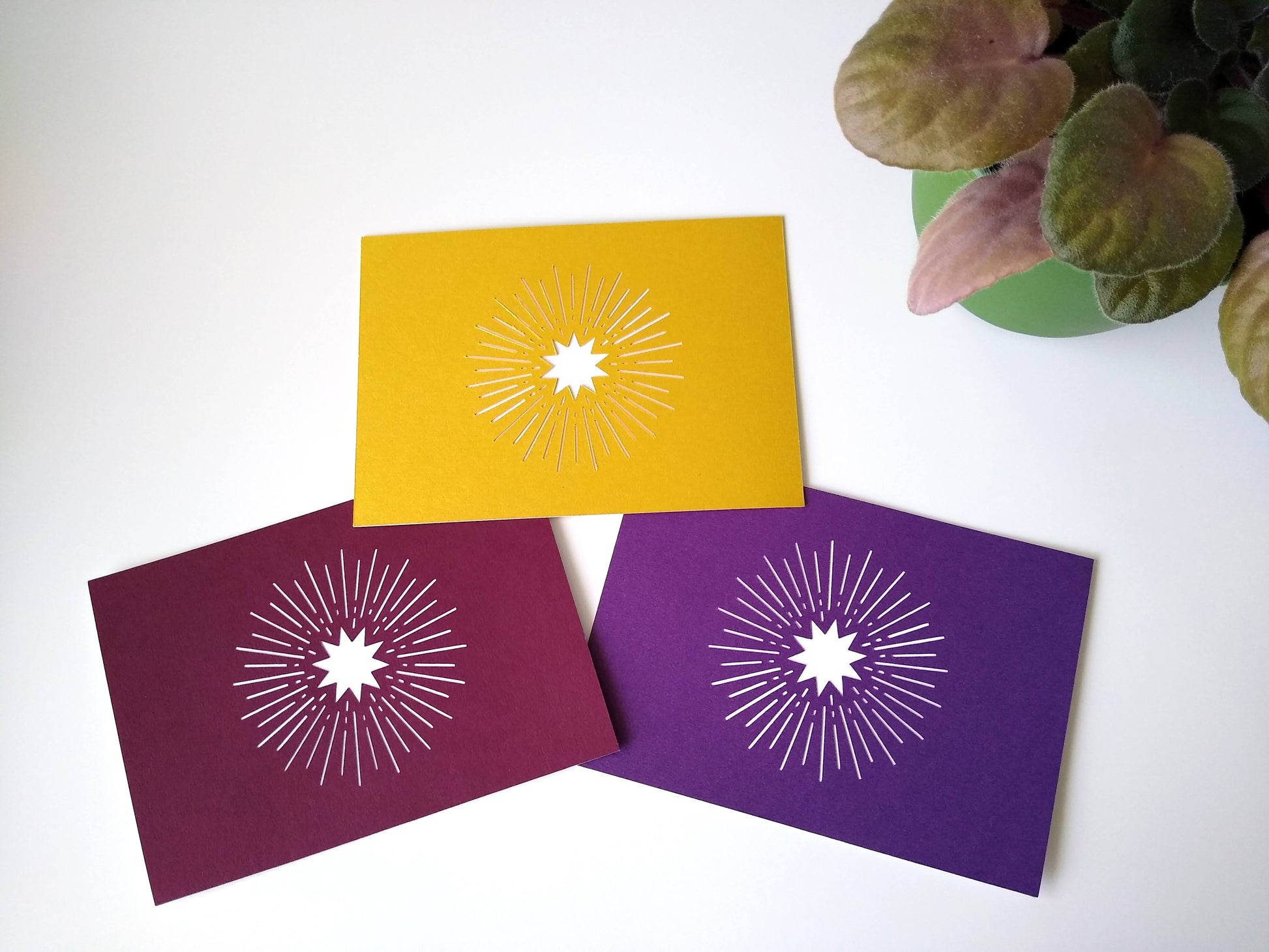 Three postcards rest on a table beside a potted plant. One is gold, one burgundy, and one metallic purple, all with a white starburst.