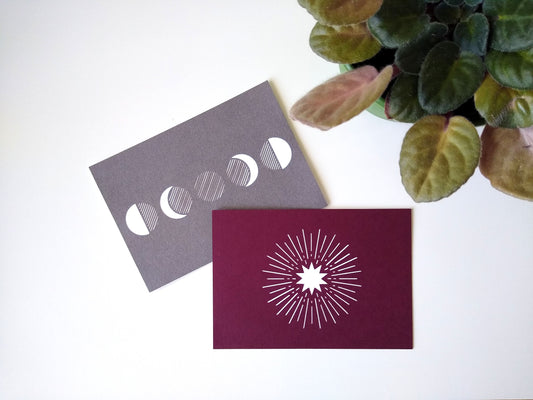 Two postcards rest on a table beside a potted plant. One is metallic grey with five moon phases on it, the other is burgundy with a white starburst.