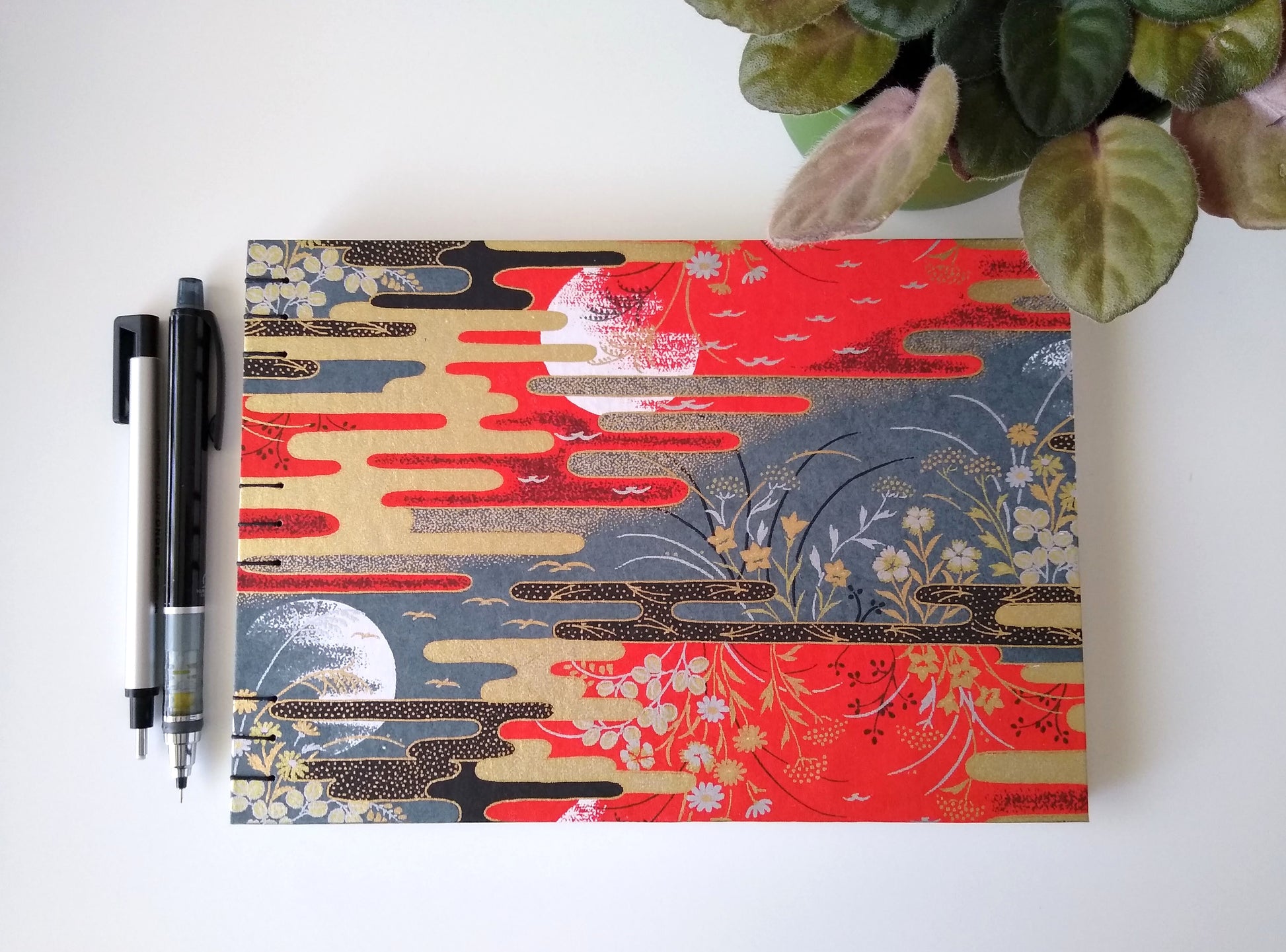 A handmade journal laying on a white desk beside a potted plant, a black mechanical pencil and a mechanical eraser. The cover of the journal is a whole scene with red and grey skies with partial moon outlines, flowers, birds, and gold and black stylized clouds.