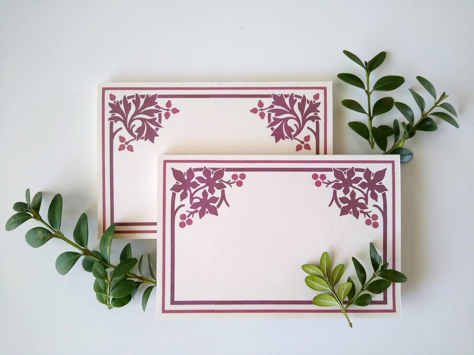 Two cream colored notepads rest on a white background. Sprigs of leaves rest around them. Each notepad has a stylized fall leaf design in burgundy in both upper corners with a double line border around the rest of the notepad.