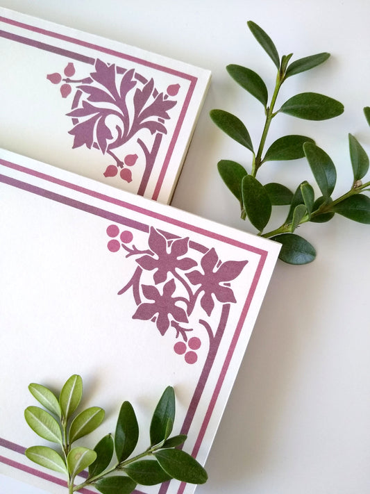 Close up of two cream colored notepads on a white background. Sprigs of leaves rest around them. Each notepad has a stylized leaf design in burgundy in the upper right corner with a double line border around the rest of the notepad.