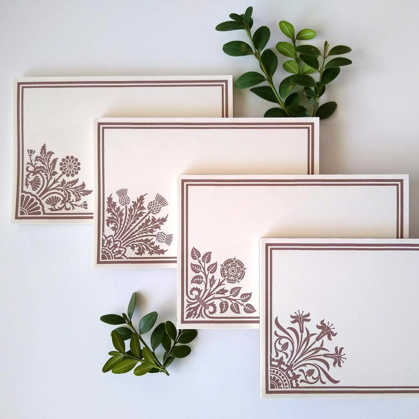 Four cream colored notepads on a white background. Sprigs of leaves rest around them. Each notepad has a stylized floral design in tan in the lower left corner with a double line border around the rest of the notepad.