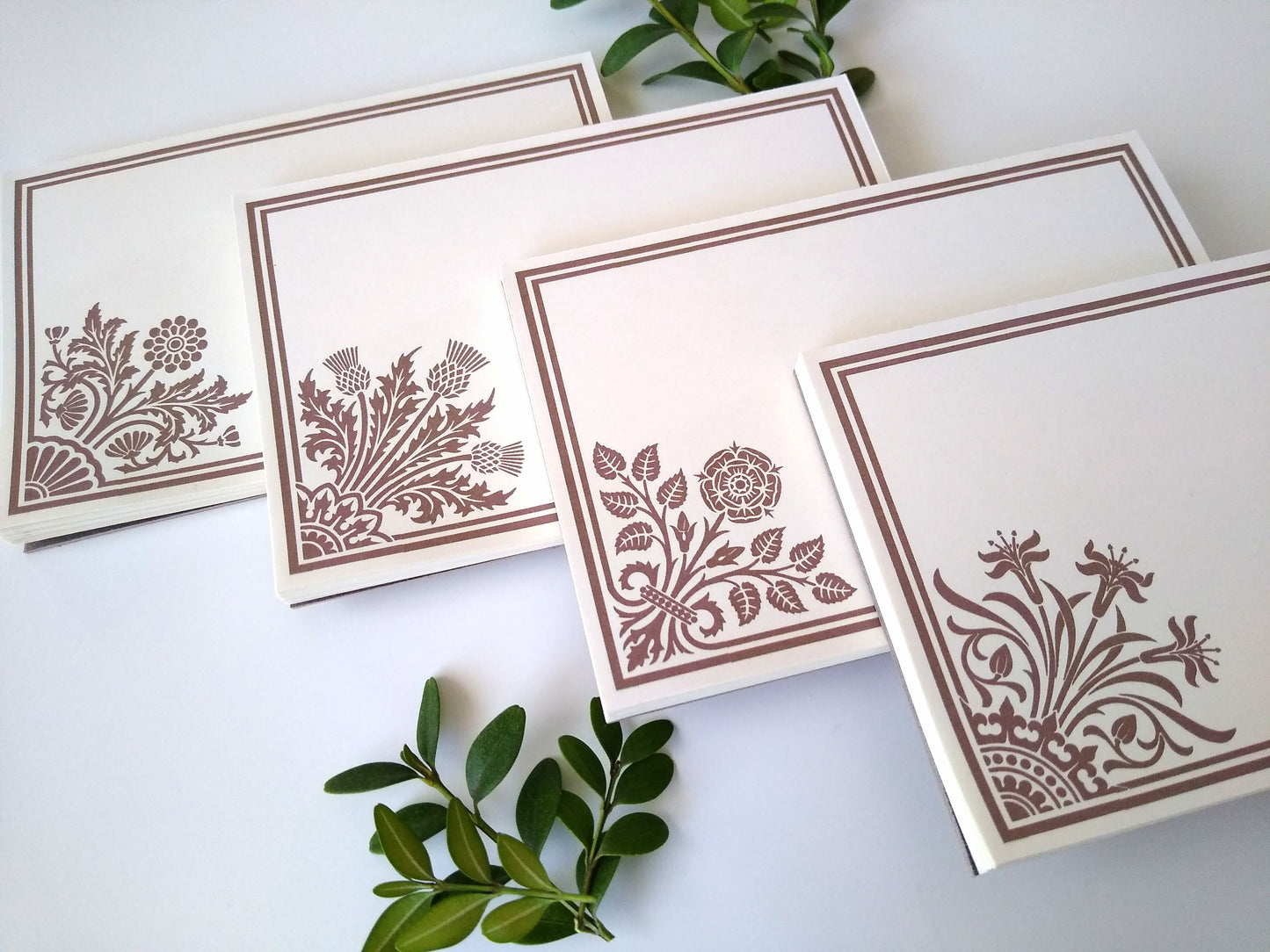 Four cream colored notepads at an angle on a white background. Sprigs of leaves rest around them. Each notepad has a stylized floral design in tan in the lower left corner with a double line border around the rest of the notepad.