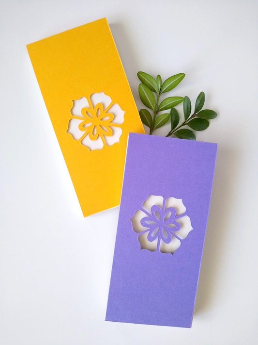 Two small notepads rest on a white background. One has a yellow cover, the other a purple cover. There are two sprigs of green leaves next to them. Cut into the center of the covers is a single hexagonal flower.
