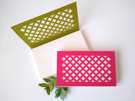 Two small notepads rest on a white background. One, with a green cover, is open. The other, with a pink cover, is closed. There are two sprigs of green leaves next to them. Cut into the covers is a lattice pattern, repeated across the entire cover.