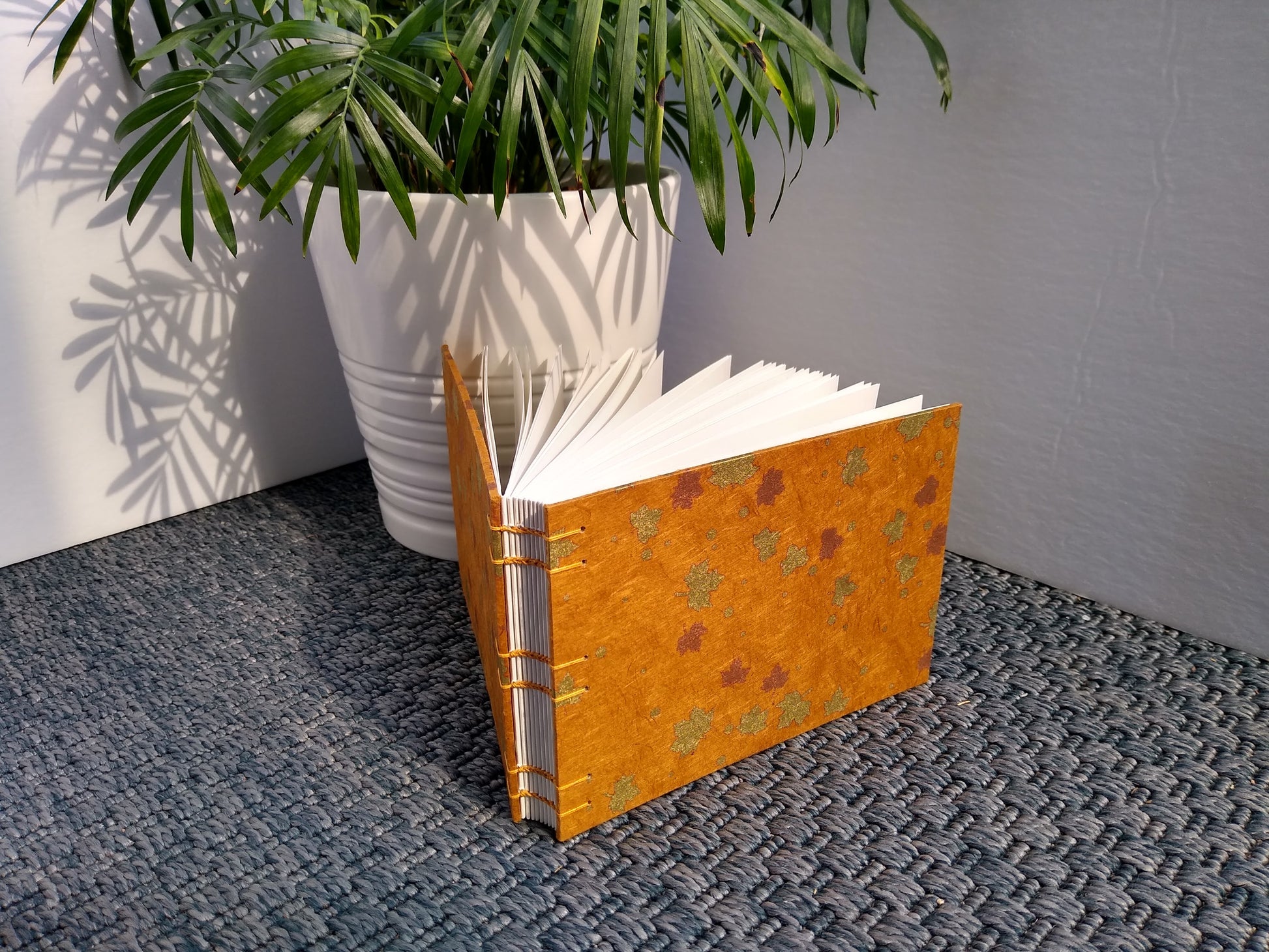 A handmade journal sits upright beside a potted plant. The cover of the journal is burnt orange with gold and copper maple leaves. It is angled to show the open spine held together with orange stitching.