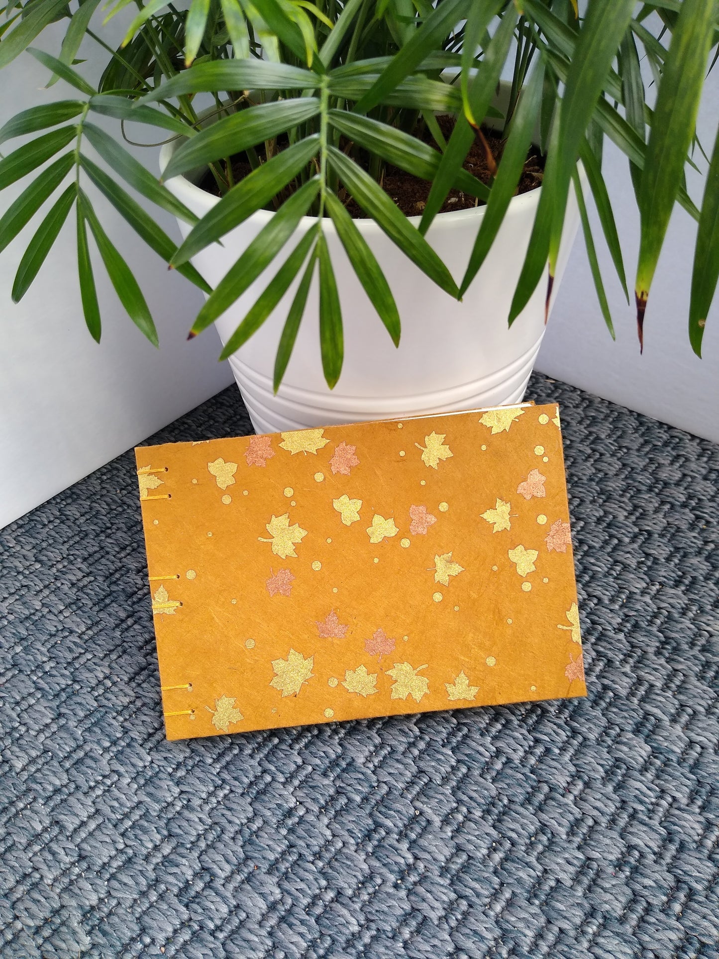 A handmade journal rests against a potted plant on a grey carpet. The cover of the journal is burnt orange with gold and copper maple leaves.