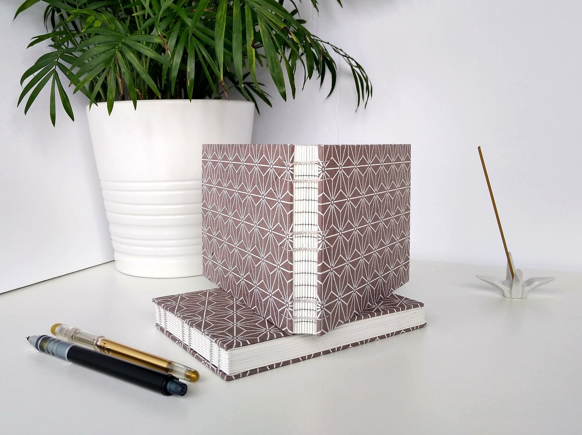 Two handmade journals are positioned beside a potted plant, a ceramic crane, a gold pen, and a black mechanical pencil. One is laying flat and the other is standing upright on top of it. The cover of the journals are grey with a white asanoha pattern repeated over its entirety. They are angled to show their open spines, stitched together with grey thread.
