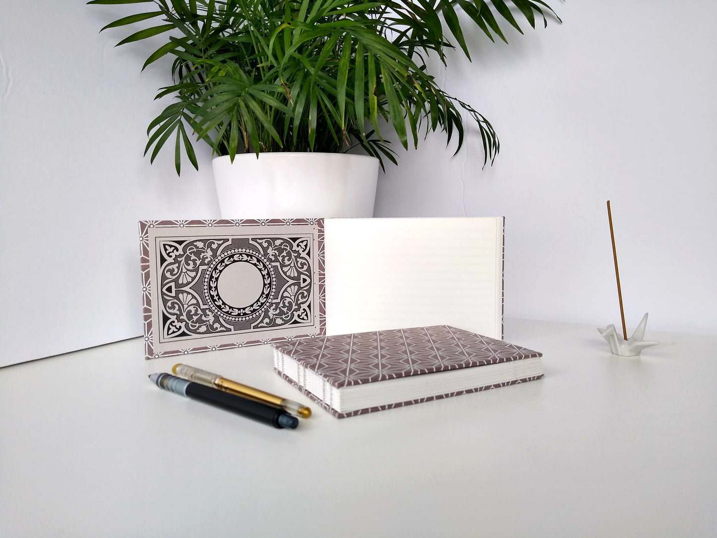 Two handmade journals are positioned beside a potted plant, a ceramic crane, a gold pen, and a black mechanical pencil. One is laying flat and the other is standing upright, opened. It shows the inside endpaper, with an intricate floral and geometric bookplate and white pages. The cover of the journals are grey with a white asanoha pattern repeated over their entirety.