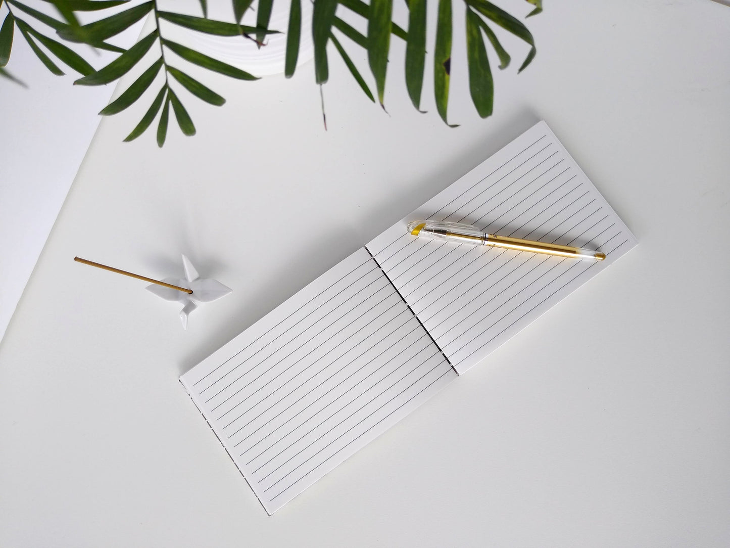 An open journal lays on a white background next to a potted plant and a ceramic crane. The journal has a grey thread connecting the pages at the spine and a gold pen rests across the lined white pages.