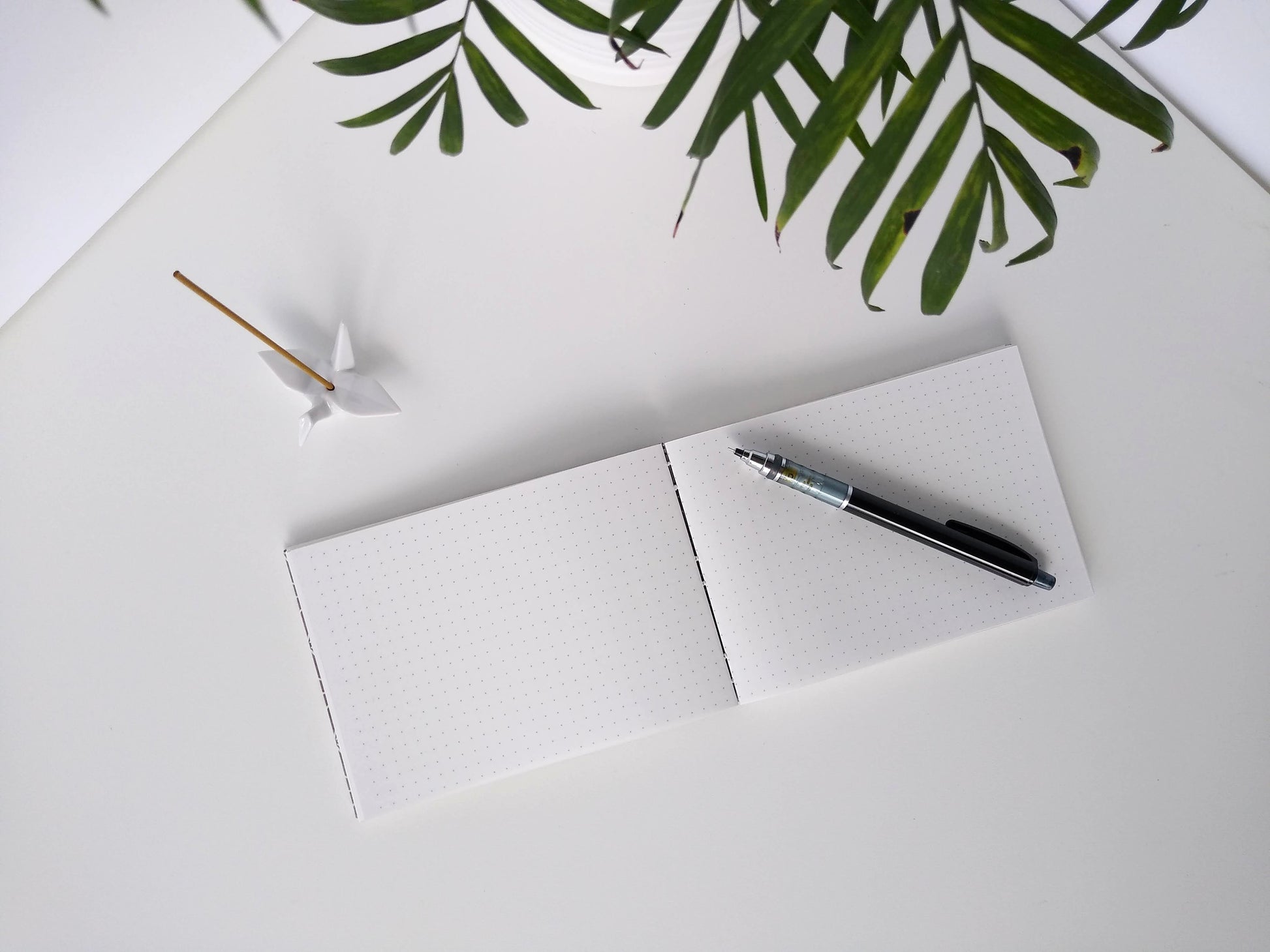 An open journal lays on a white background next to a potted plant and a ceramic crane. The journal has a grey thread connecting the pages at the spine and a black mechanical pencil rests across the white pages with a printed dot grid.