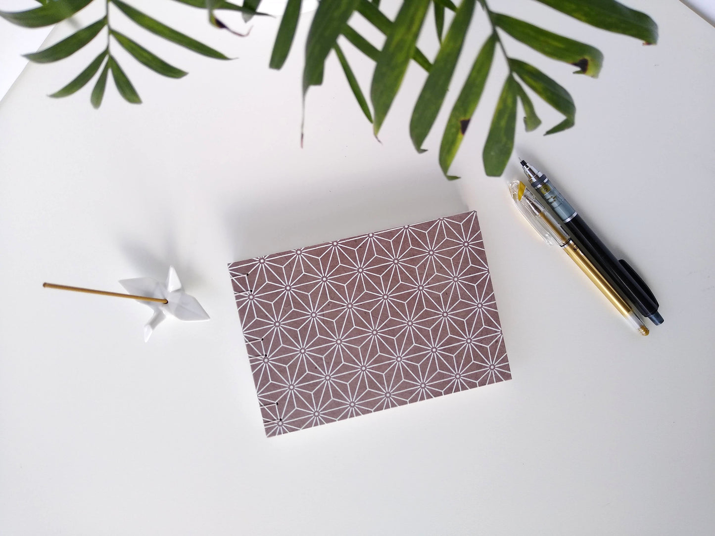 A handmade journal laying on a white desk beside a potted plant, a ceramic crane, a gold pen, and a black mechanical pencil. The cover of the journal is grey with a white asanoha pattern repeated over its entirety.
