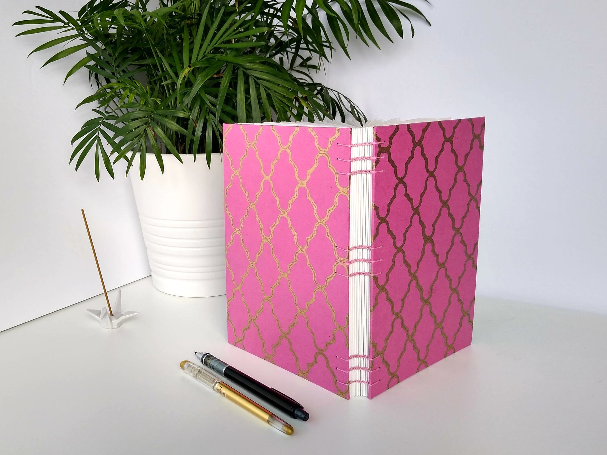 A handmade journal sits upright beside a potted plant, a ceramic crane, a gold pen, and a black mechanical pencil. The cover of the journal is pink with a gold quatrefoill design across the front. The journal is turned to show the open spine with pink stitching to hold it together.