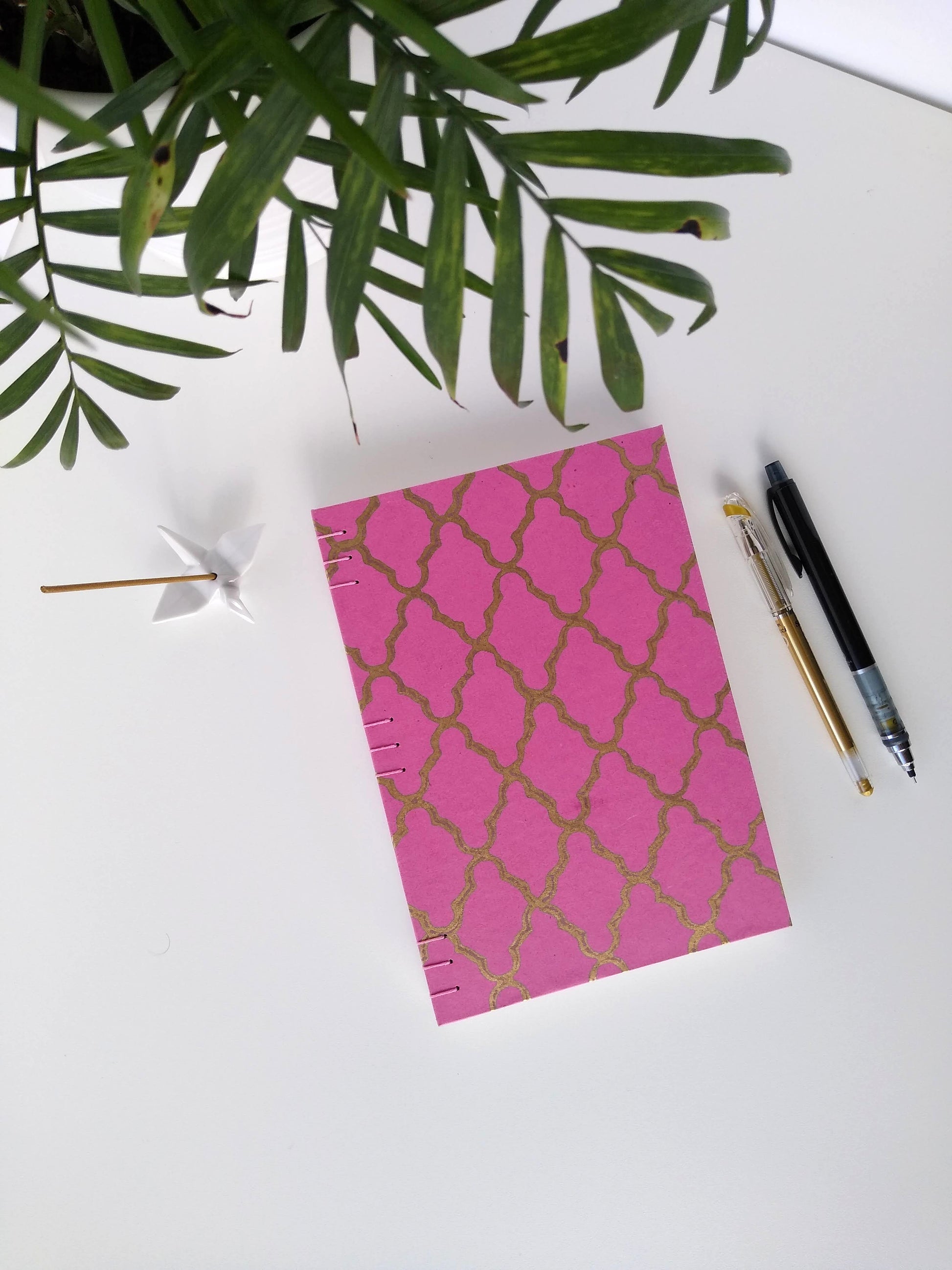 A handmade journal laying on a white desk beside a potted plant, a ceramic crane, a gold pen, and a black mechanical pencil. The cover of the journal is pink with a gold quatrefoill design across the front.