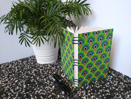 A handmade journal sits upright beside a potted plant, a black pen, and a black mechanical pencil. The cover of the journal is an abstract peacock feather design. It's angled to show the open spine and the black thread stitching it together.